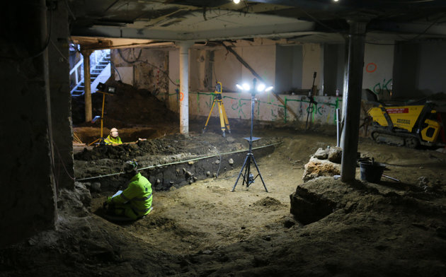 Foto af udgravningen i København, hvor arkæologer badet i projektørlys udgraver Clemensgrøften i kælderen af den eksisterende bebyggelse