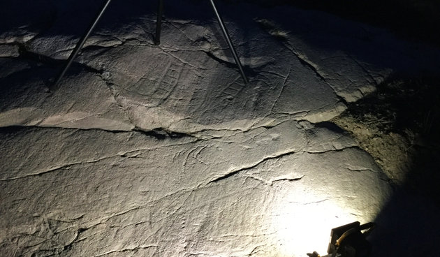 Foto af kræftig lyskilde, der kaster et lys langs klippens overflade og fremhæver skyggerne af de indridsede skibe fra bronzealderen.