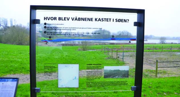 På den opstillede glasvæg kan du se helt præcist, hvor i søen man fandt de fire store våbenofringer. Foto: Kulturstyrelsen Foto af informationstavlen, der er ne glasvæg, så man helt præcist kan se, hvor i søen man fandt de fire store våbenofringer