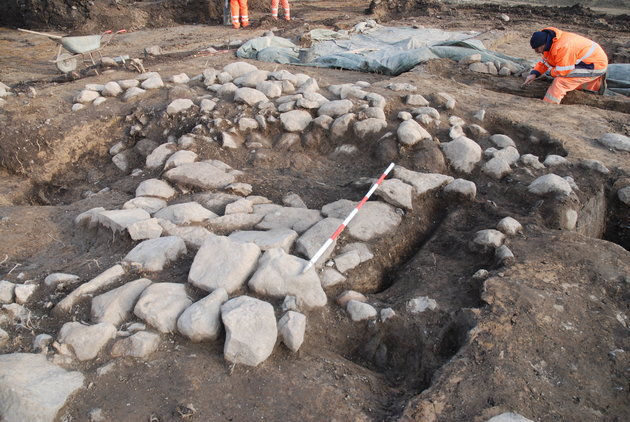 Billede fra udgravningen, hvor stenpakningen er ved at blive renset af for jord af arkæolog i arbejdstøj.