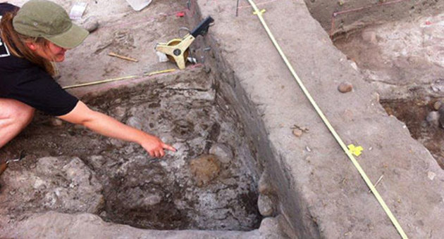 Foto af arkæolog, der peger på skjoldbulen i en våbengrav fra ældre jernalder, der netop er kommet til syne i udgravningen.