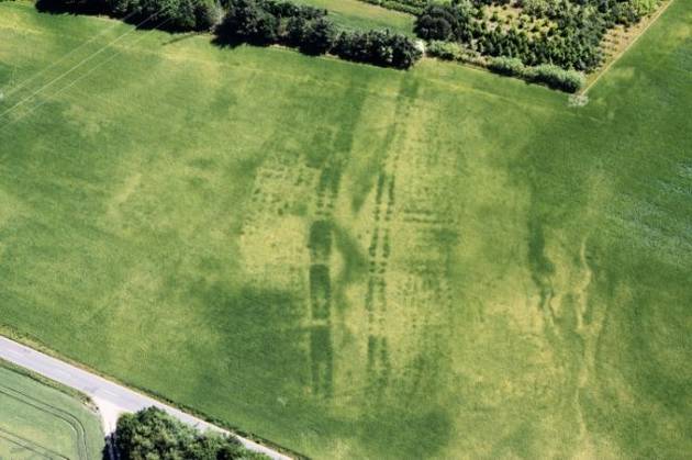 Fra luften ses sporene efter Jernalderens huse. Foto: Lis Helles Olesen Fra luften ses en mark, hvor afgrøderne står med forskellige farvetoner. Farvetonerne er spor efter flere huse fra jernalderen, der ligger på lange rækker.