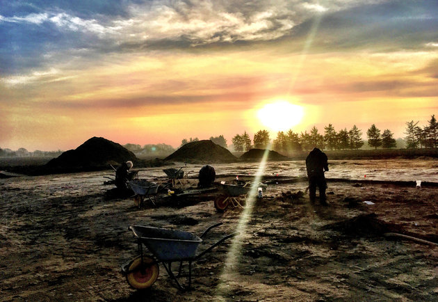 Solnedgang over den arkæologiske udgravning, hvor arbejdende arkæologer fremstår som sorte siluetter mod den orange himmel