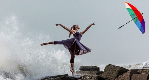 Kvindelig balletdanser danser på klipper med høje bølger i baggrunden og en paraply,der er ved at blæse væk.