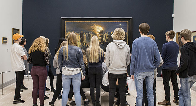 En gruppe af unge står foran Sankt Hansblus på Skagen Strand malet af Michael Ancher.